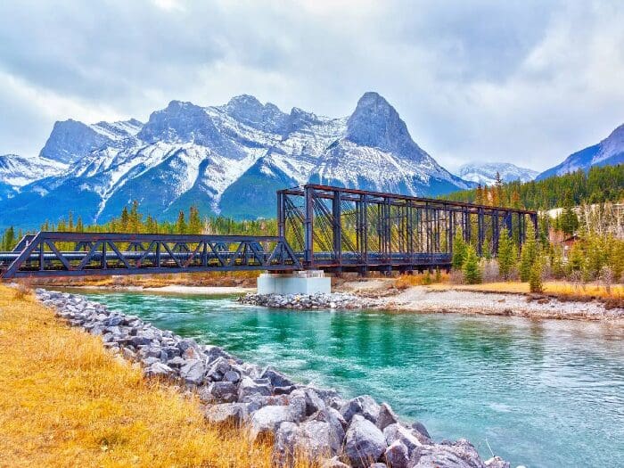 Metal bridge over river with snowy mountains.